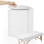Stack of white disposable non-woven fabric massage table sheets, unfolded on a treatment bed.