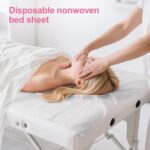Stack of white disposable non-woven fabric massage table sheets, unfolded on a treatment bed.